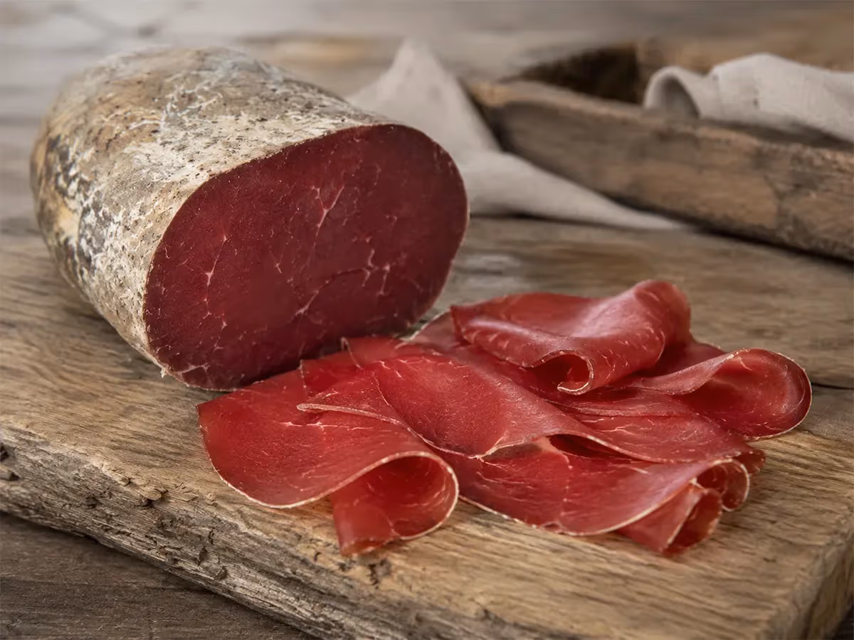 Pezzo intero di bresaola con crosta speziata affiancato da fette rosse su tagliere di legno antico