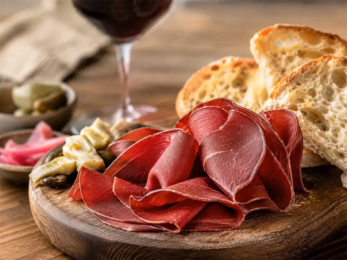 Fettine di bresaola arrotolate su tagliere rotondo con pane croccante olive capperi e calice di vino rosso