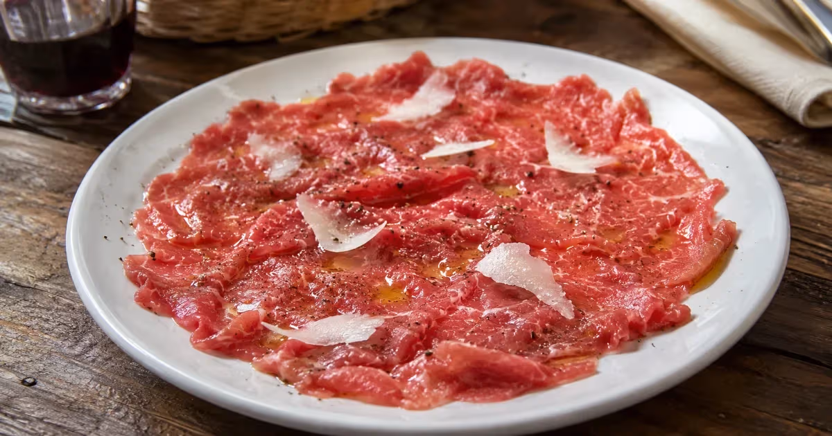Carne cruda all'albese su piatto bianco con scaglie di parmigiano olio e pepe nero su tavolo rustico in legno