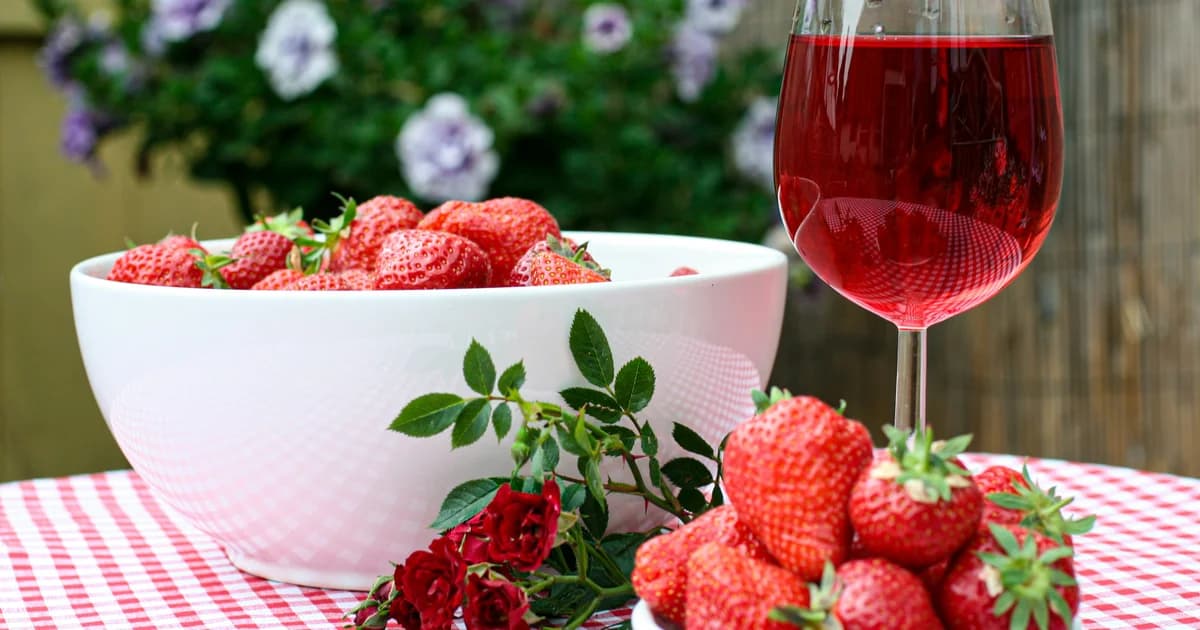 Ciotola bianca colma di fragole fresche rosse accanto a calice di vino rosso su tovaglia a quadretti
