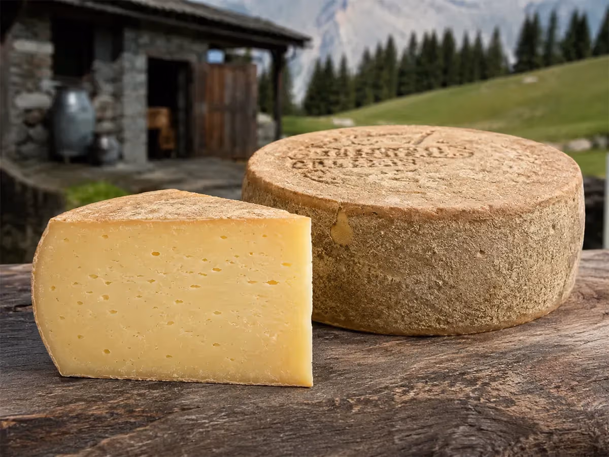 Forma intera e spicchio di Montasio DOP su tronco di legno con malghe alpine sullo sfondo