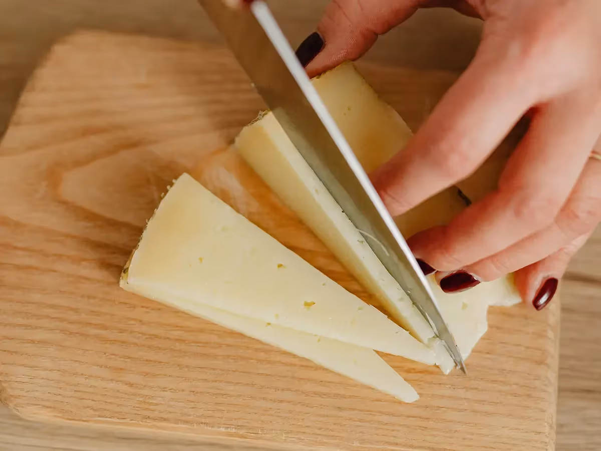 Mani femminili che affettano formaggio a pasta gialla paglierina con coltello su tagliere chiaro