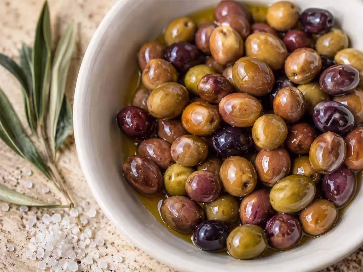 Olive taggiasche lucide verdi e viola in ciotola bianca con sale grosso e ramoscello d'ulivo