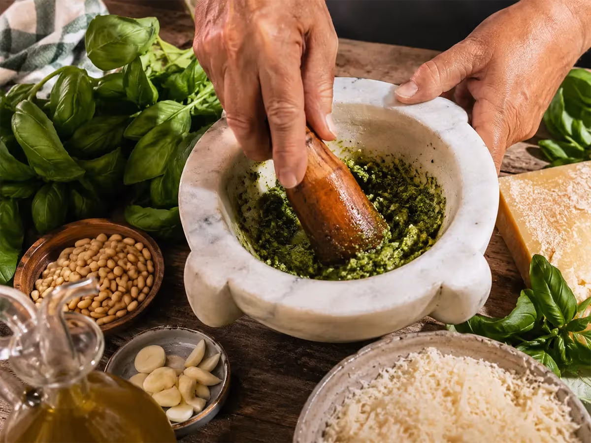Mani che pestano pesto alla genovese verde brillante in mortaio di marmo con basilico fresco e pinoli
