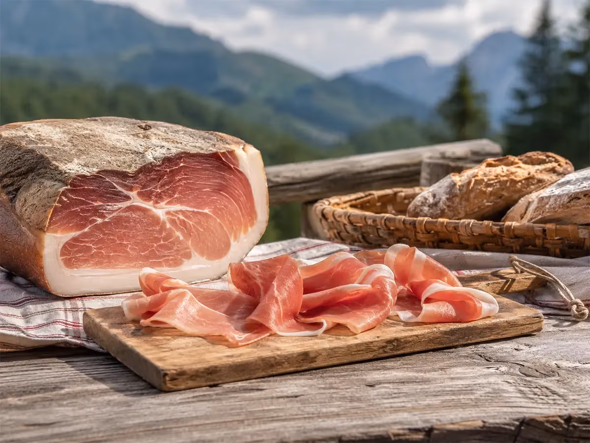 Coscia di prosciutto stagionata con fette rosa su tagliere e cesto di pane con montagne sullo sfondo