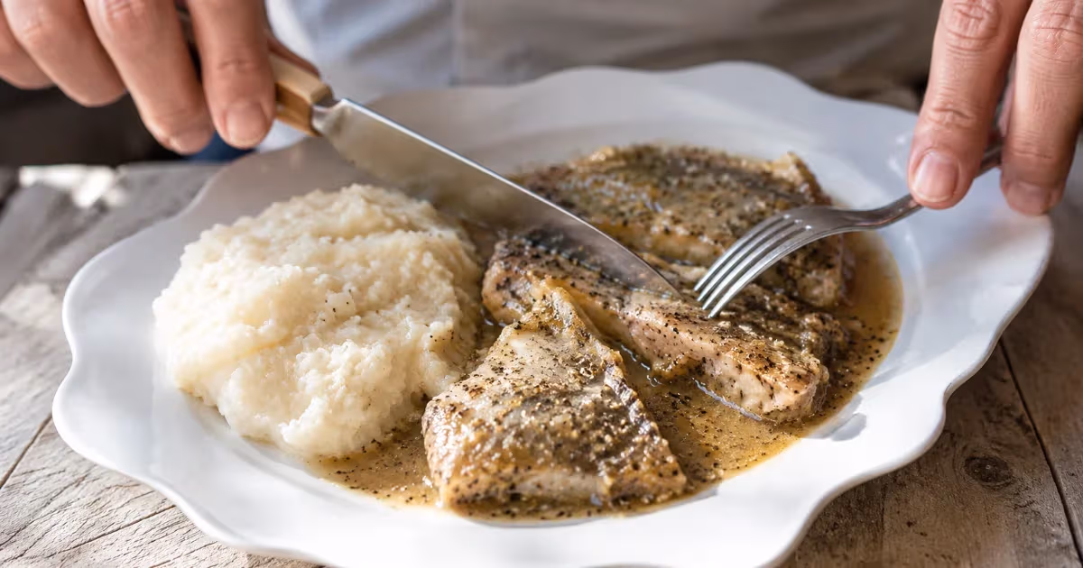 Boreto alla gradese con filetti di pesce in salsa scura su piatto bianco smerlato accanto a polenta bianca cremosa tagliato con coltello e forchetta